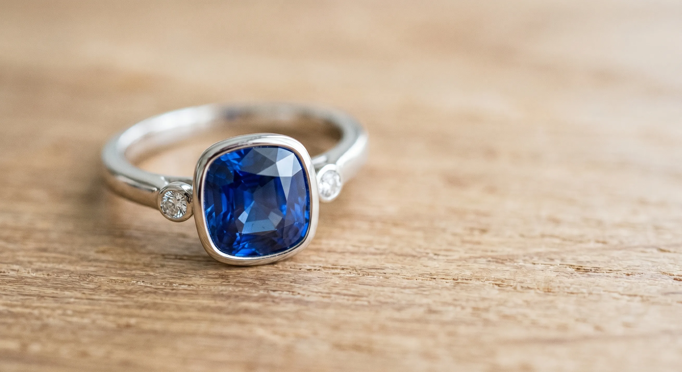 A Ceylon sapphire engagement ring photographed from above on a softly lit warm wood surface, showing a continuous bezel setting holding a cushion-cut blue sapphire securely in white gold